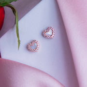 A pair of rose gold heart-shaped stud earrings with white stone detailing, displayed on a pink fabric.